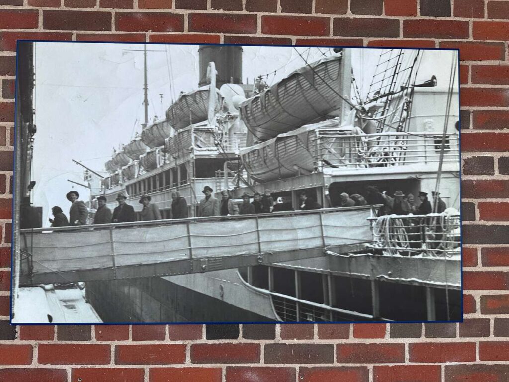 Archival photograph mounted on a brick wall showing passengers crossing a gangway from a ship at port, with lifeboats and deck structures visible above them.