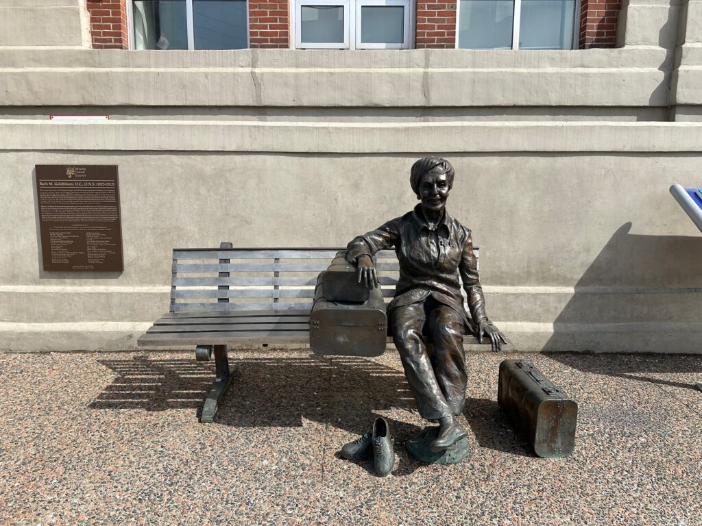 Bronze statue of Ruth M. Goldbloom seated beside vintage suitcases on a bench outside Pier 21, with a commemorative plaque mounted on the wall behind her.