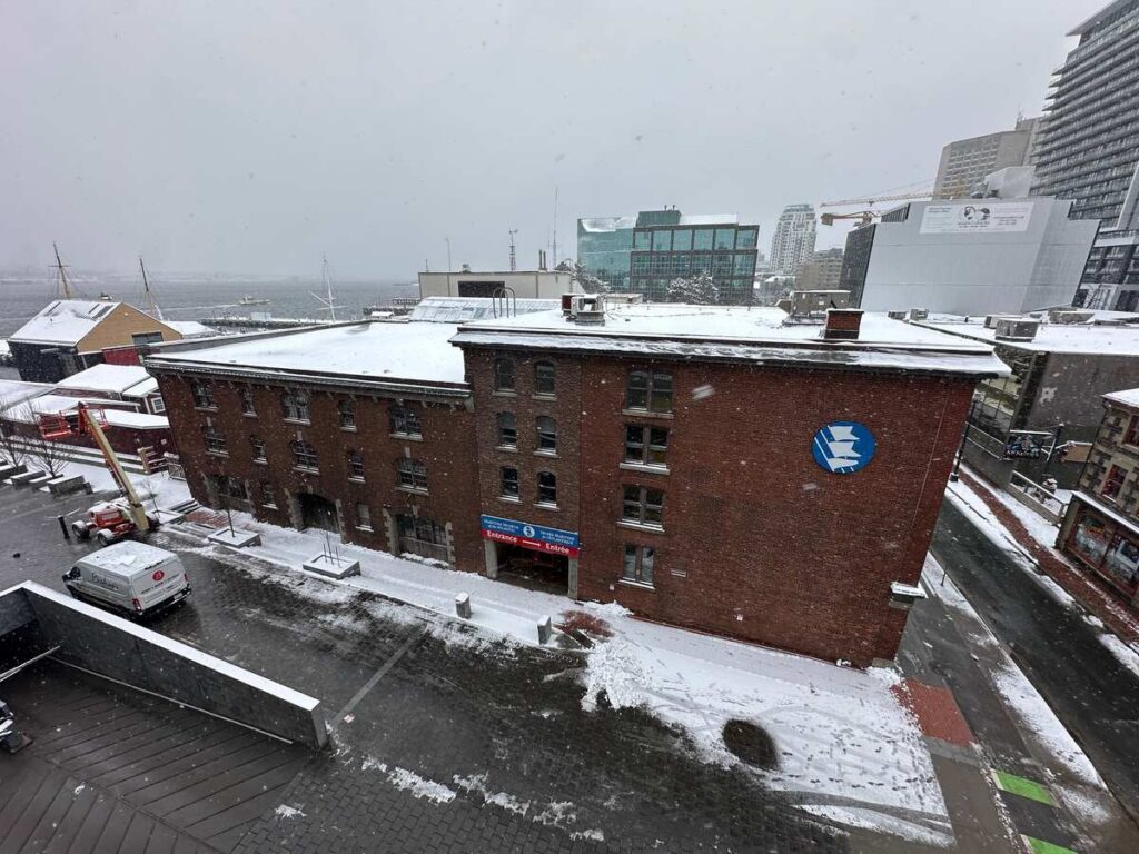 Snow falls over the brick exterior of the Maritime Museum of the Atlantic in Halifax, with Halifax Harbour visible in the background.
