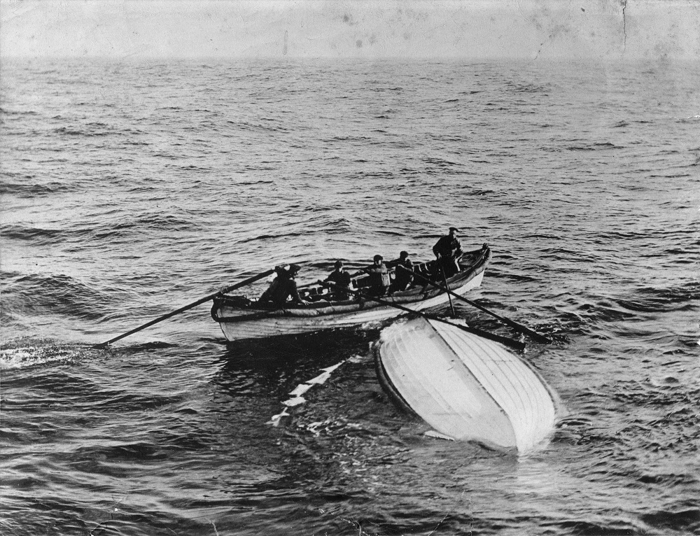 Black-and-white historic photograph showing a small lifeboat beside an overturned boat on open water during the Titanic recovery effort.