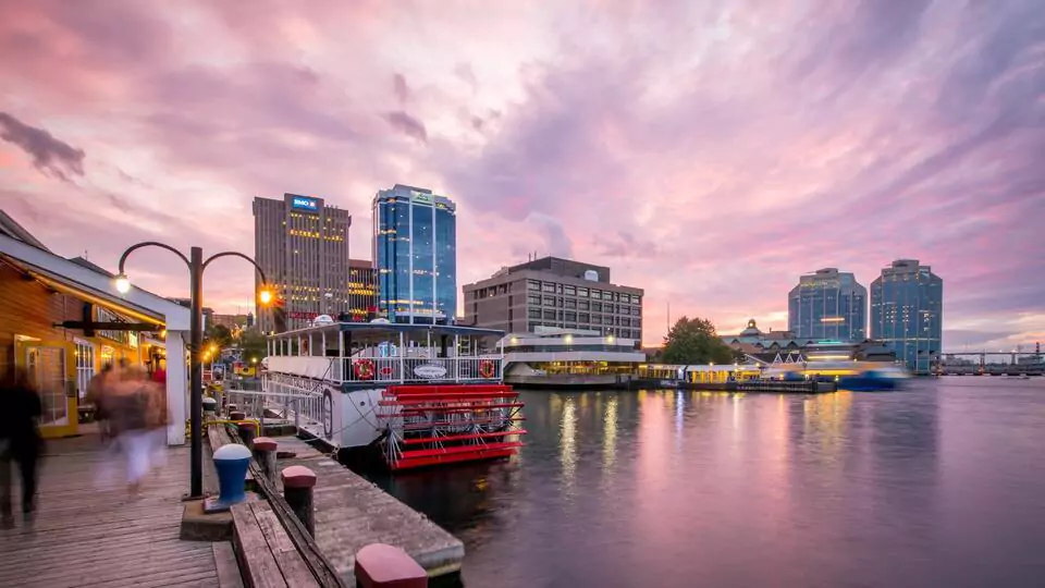 Halifax waterfront at sunset with boardwalk, paddle boat, and glowing skyline reflections.