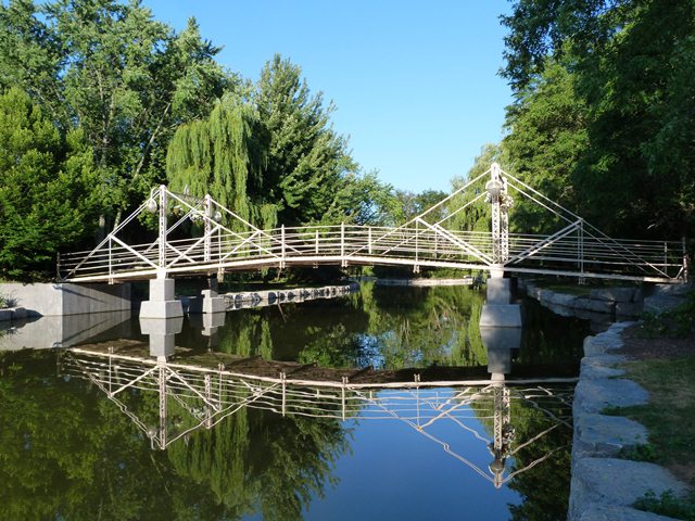 White Victorian-style pedestrian swing bridge reflected in calm water in Lindsay Ontario