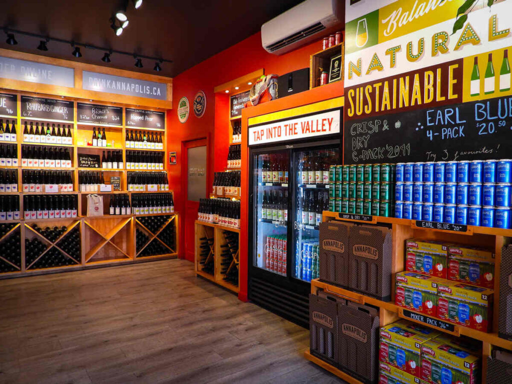 Brightly coloured craft beverage shop interior with shelves of bottled cider, canned drinks, and a fridge under a sign reading “Tap Into the Valley.”