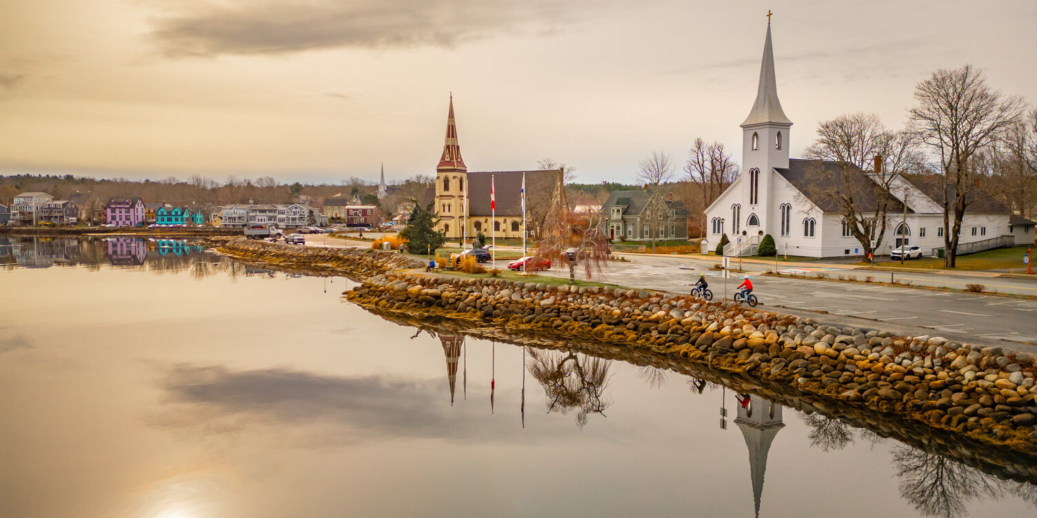 A calm view of Mahone Bay’s waterfront at golden hour, with two of the town’s iconic churches reflected in still water, a stone shoreline in the foreground, and a soft peach sky stretching above the small coastal community.