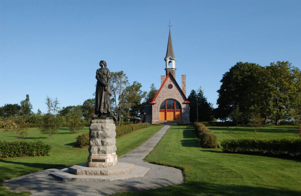 Statue of Evangeline standing on the lawn at Grand-Pré, with the memorial church and a clear blue sky behind her.