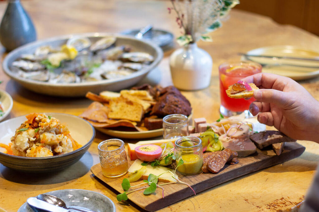 Table spread with local seafood, oysters, charcuterie, pickles, bread, and cocktails, with a hand lifting a canapé.