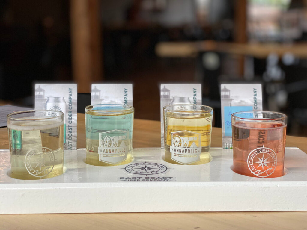 Four small tasting glasses of cider in different colours arranged in a flight on a wooden bar.