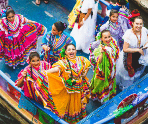 A group of smiling performers in bright multicoloured traditional dresses dance aboard a decorated river barge during a previous Fiesta San Antonio celebration.