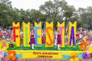 A colourful Fiesta San Antonio Commission parade float featuring giant floral letters spelling “FIESTA” moves past cheering crowds during the celebration.