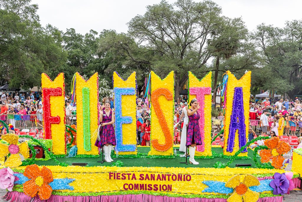 A colourful Fiesta San Antonio Commission parade float featuring giant floral letters spelling “FIESTA” moves past cheering crowds during the celebration.