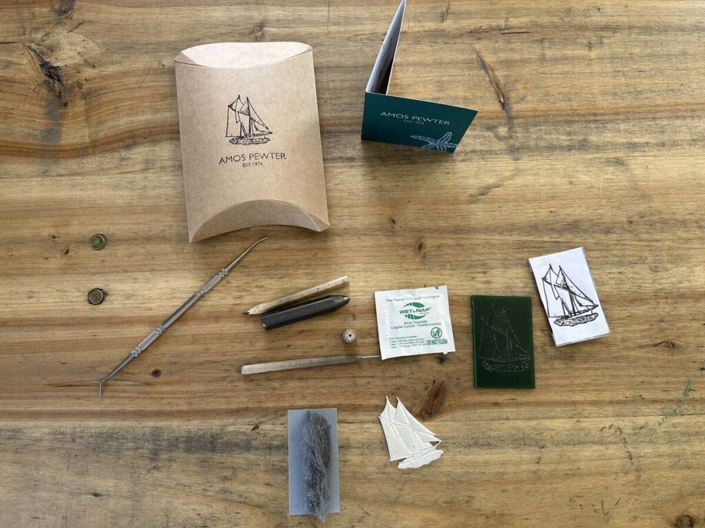 Wax-carving tools and materials laid out on a wooden table at Amos Pewter, including carving picks, sanding wool, mould pieces, and a Bluenose design template.