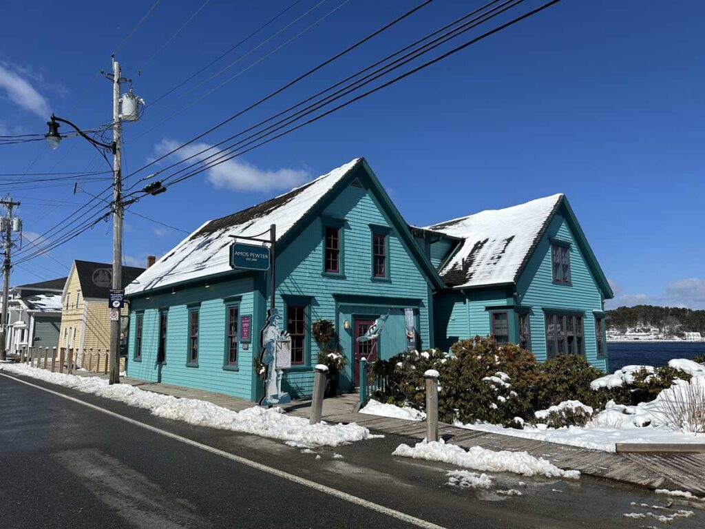 Exterior of Amos Pewter in Mahone Bay, a turquoise waterfront heritage building with snow on the roof and bright blue sky overhead.