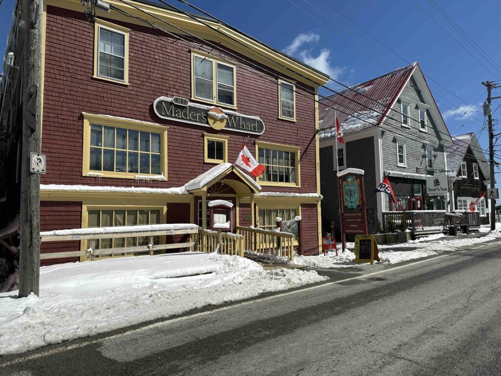 Snow-lined street in Lunenburg with the historic Old Mader’s Wharf building in deep red, Canadian flags out front, and neighbouring waterfront businesses beside it under a bright blue sky.