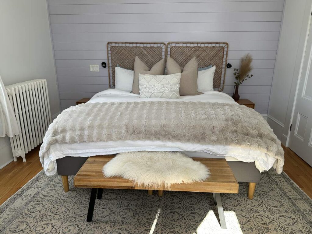 Light-filled bedroom at The Ivy House Inn with a large bed, layered neutral textiles, woven headboard panels, and a white radiator by the window.