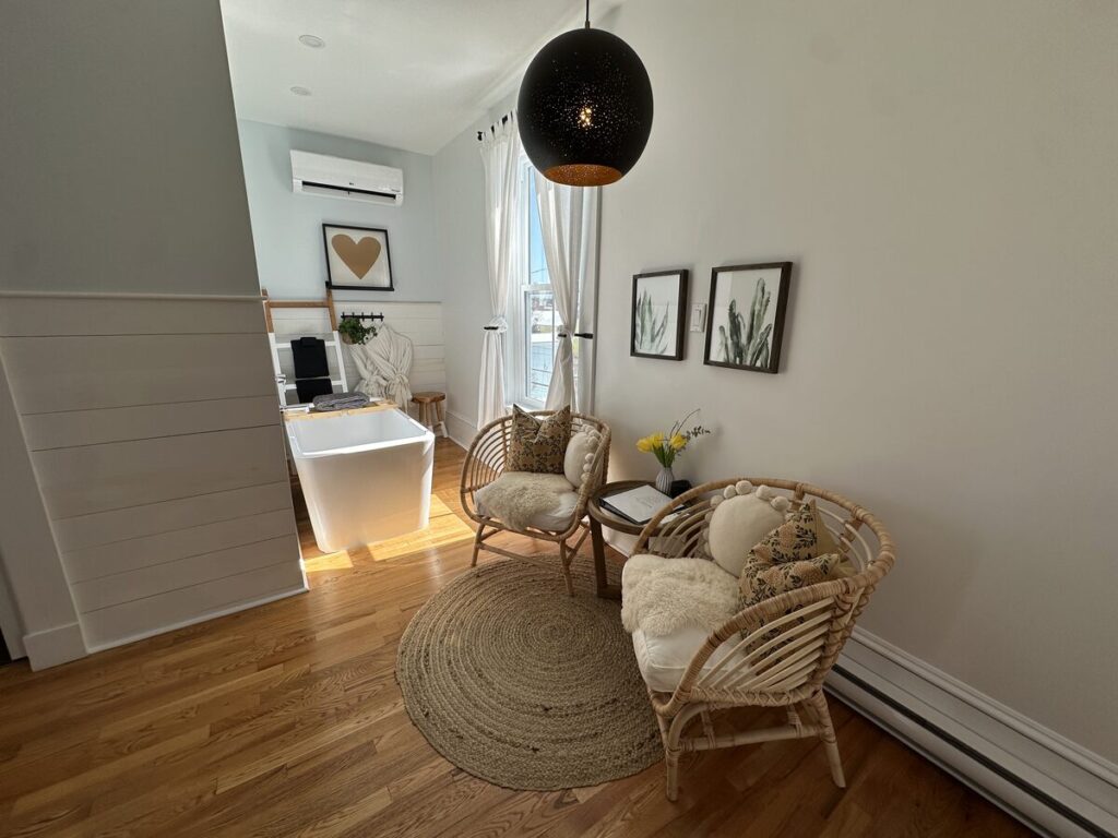 Sitting area inside a suite at The Ivy House Inn with two wicker chairs, a round jute rug, tall windows, and a freestanding bathtub in the background.