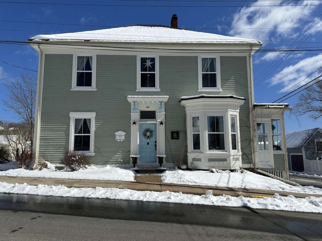 Exterior of The Ivy House Inn in Lunenburg, a pale green heritage home with white trim, a light blue front door, and snow along the sidewalk.