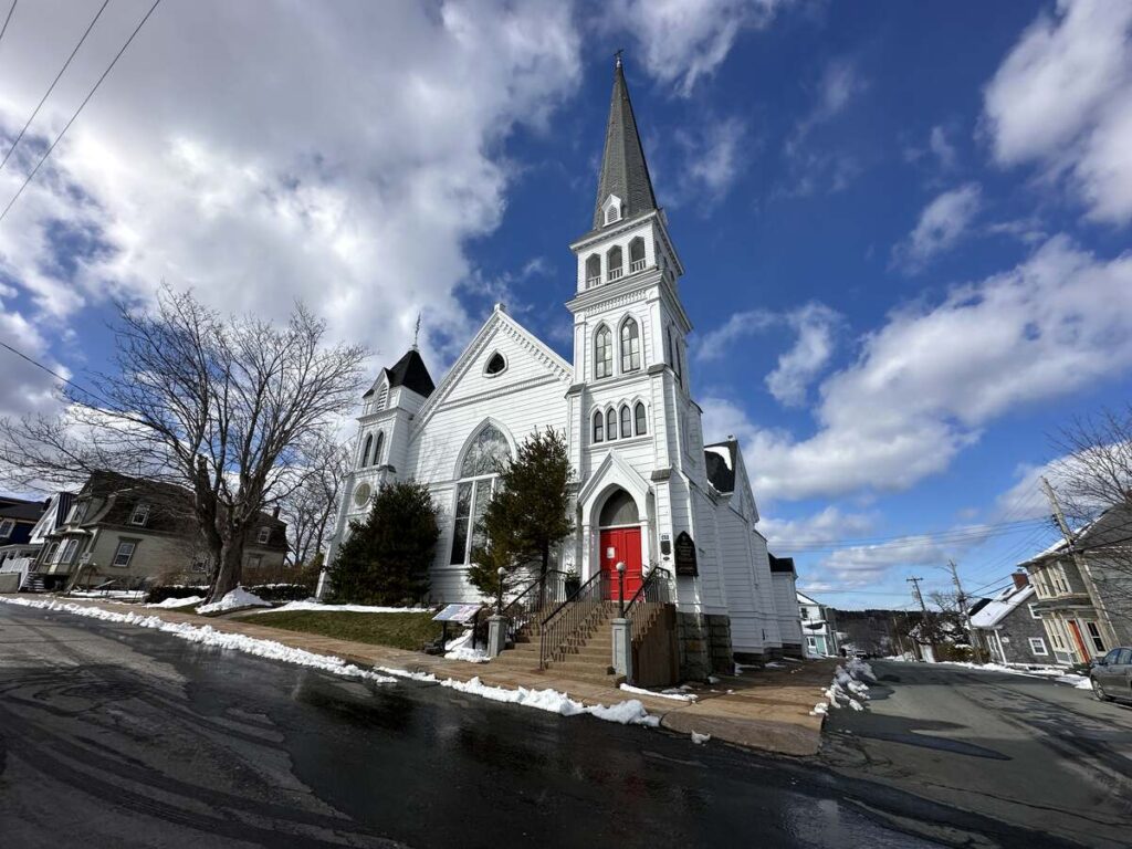 Exterior of St. John’s Anglican Church in Lunenburg, a white wooden church with twin towers, a tall central spire, and bright red doors.