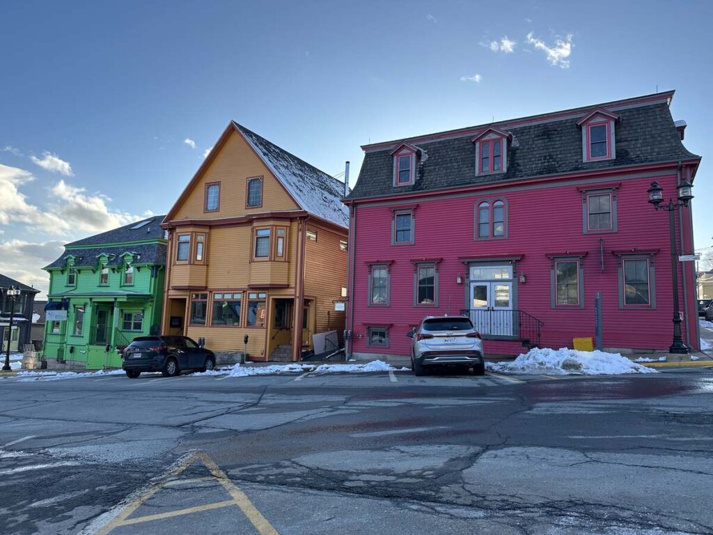 Row of brightly painted heritage buildings in Lunenburg, including pink, orange, and green facades, with snow on the ground and afternoon light on the street.