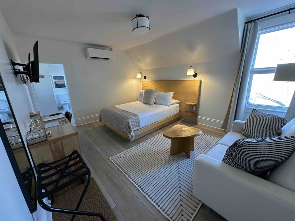 A minimalist guest room with a wooden bed, white linens, a small sofa, a desk, and soft natural light coming through two windows.