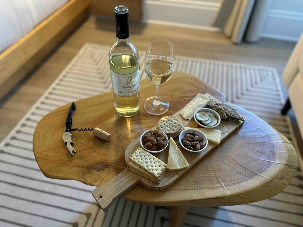 A bottle of Tidal Bay wine and a filled glass sit beside a cheese board with crackers, nuts, preserves, and pickles on a small wooden table.