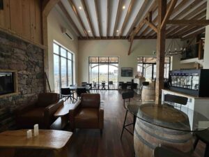 A warm, rustic-modern winery tasting room with exposed wooden beams, a stone fireplace, leather armchairs, small tables, and large windows overlooking the vineyard.