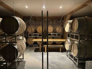 Oak barrels line both sides of a dimly lit cellar room behind glass doors, with a long wooden table at the centre and a stone wall at the back.