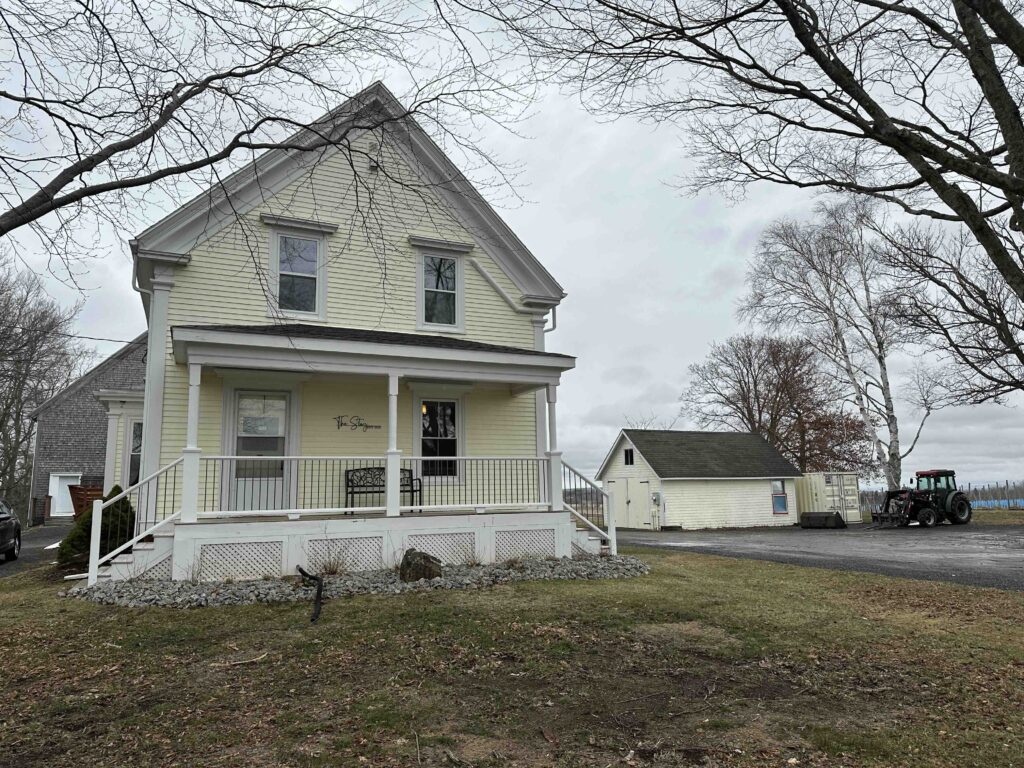 A pale yellow farmhouse-style building with a front porch sits on winery grounds, with vineyard outbuildings and a tractor nearby.