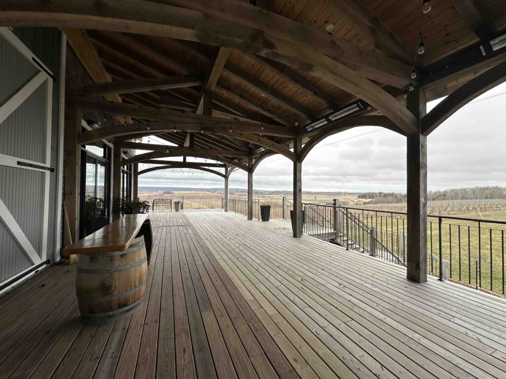 A large covered wooden deck at Planters Ridge overlooks vineyard rows and rolling valley views under a cloudy sky, with open space designed for gatherings and events.