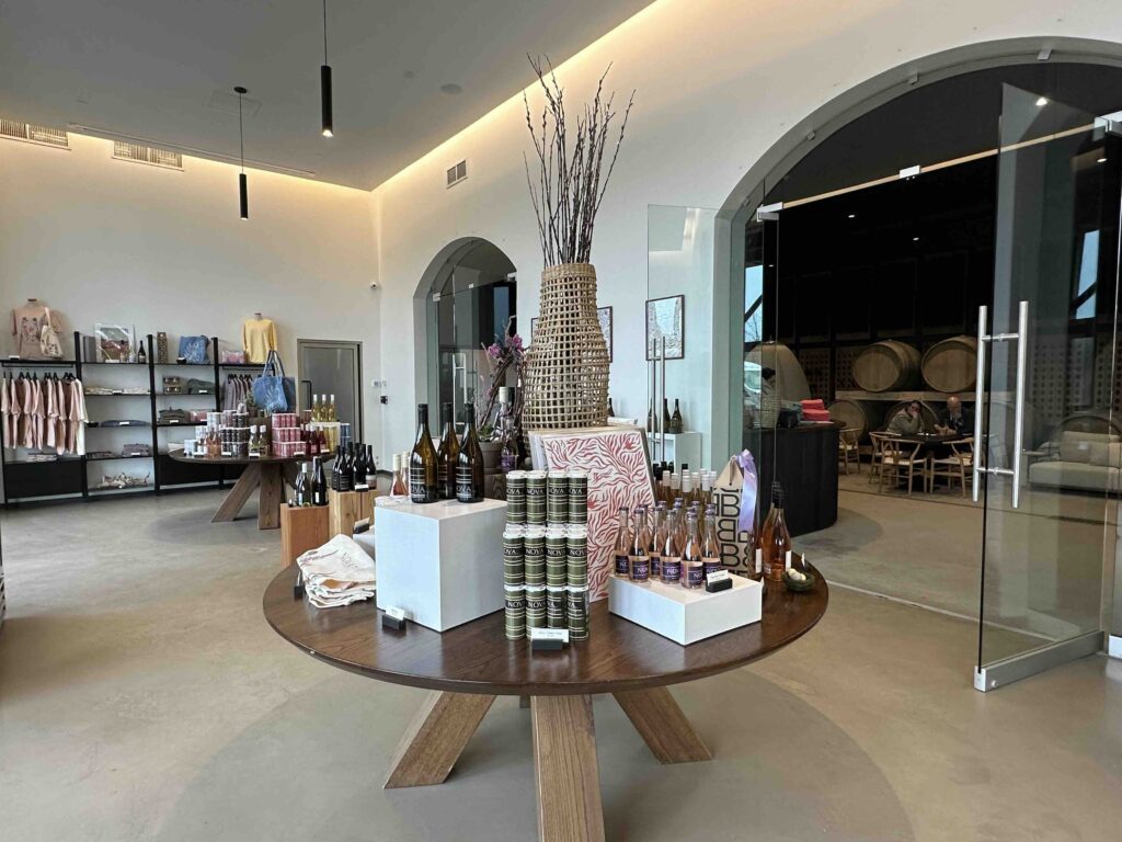 A bright, modern winery tasting room with a round display table of bottles and gifts, arched doorways, and oak barrels visible in an adjoining room.