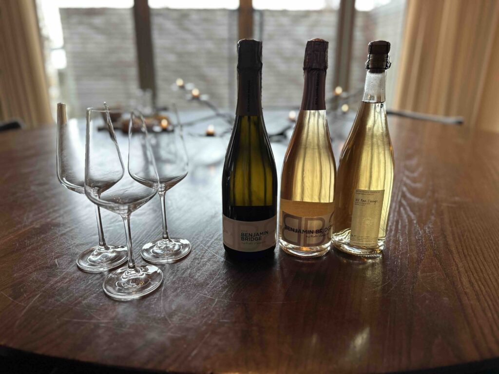 Three bottles of Benjamin Bridge wine and sparkling wine stand beside four empty glasses on a wooden tasting table, with soft light in the background.