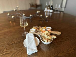 A glass of white wine sits on a wooden table beside a charcuterie board with bread, cheese, fruit, and small accompaniments in a softly lit tasting room.