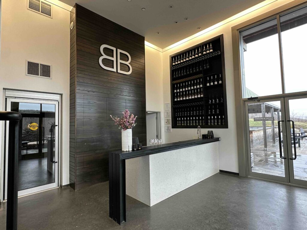 A sleek winery reception area with a large BB logo on a dark wood wall, a minimalist tasting counter, and tall windows looking outside.