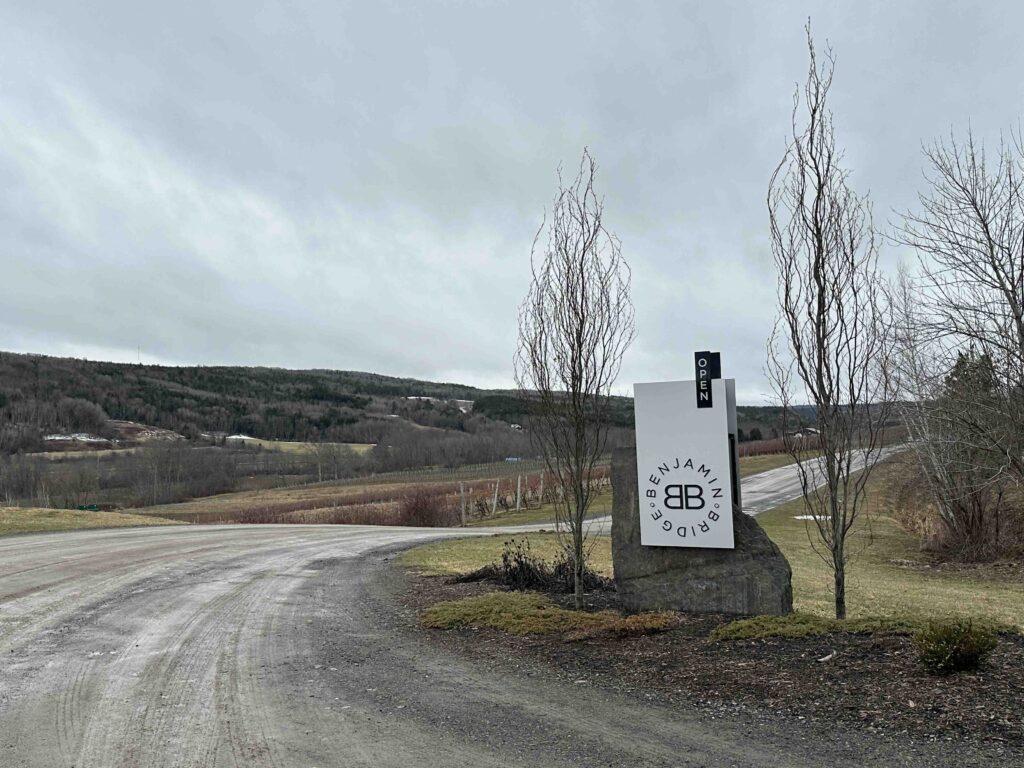 A roadside Benjamin Bridge sign stands beside a gravel lane overlooking vineyards, leafless trees, and rolling countryside beneath a cloudy sky.