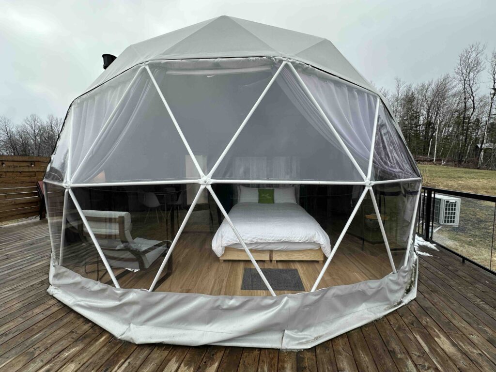 Geodesic glamping dome at Valley Sky in Nova Scotia, shown from the front on a wooden deck, with a bed, chairs, and sheer curtains visible inside under a grey sky.