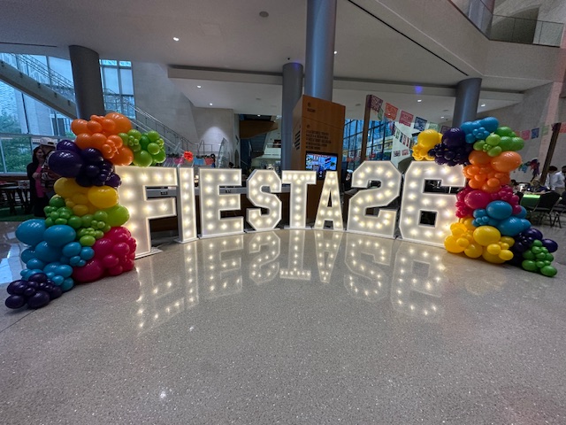 Illuminated Fiesta 26 marquee letters stand inside a modern event space, framed by bright balloon arrangements in rainbow colours.