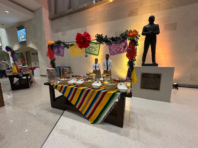 A buffet table draped in a colourful striped cloth sits beneath festive floral décor, with two staff members standing behind it near a statue inside the venue.