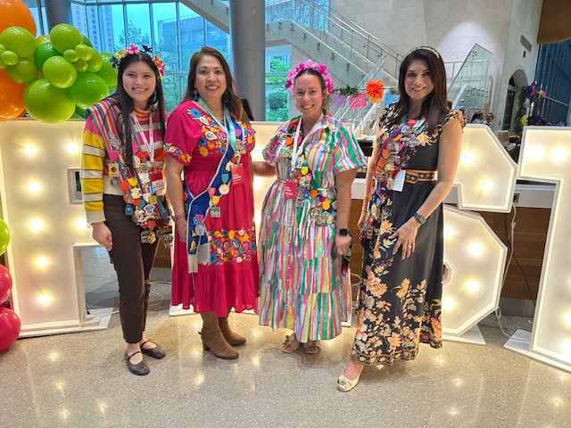 Four women pose in front of the illuminated Fiesta sign, wearing colourful outfits and medal-covered sashes at an indoor reception.