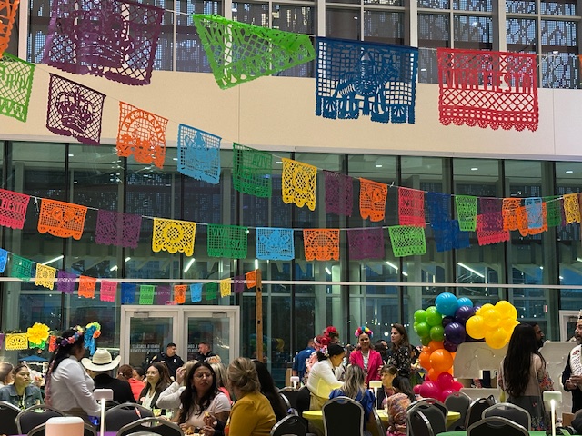 Guests gather at round tables beneath rows of colourful papel picado banners and balloon arrangements inside a large glass-walled event space.