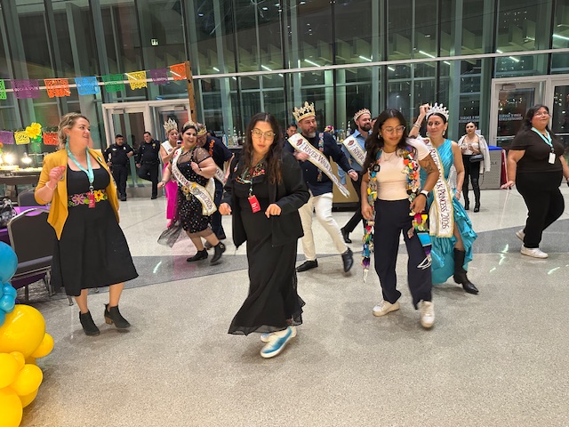 A group of guests, including Fiesta royalty and attendees wearing medals, dance together in the reception space.