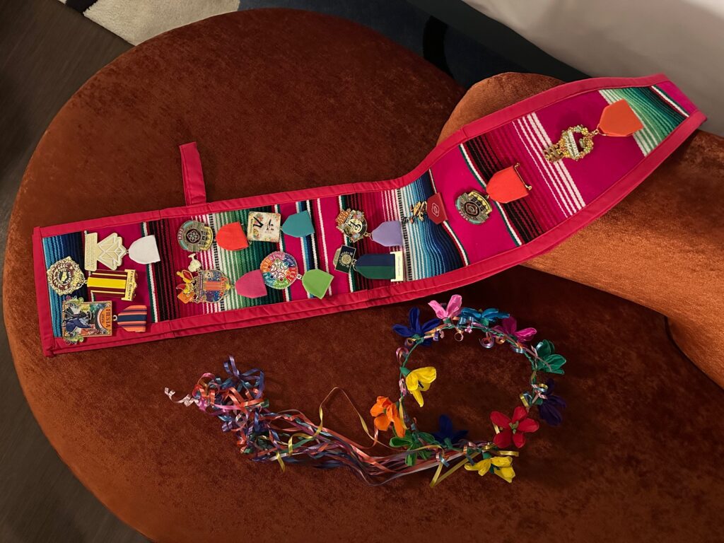 A colourful Fiesta medal sash covered in pins and ribbons is draped across a rust-coloured velvet chair, with a bright flower crown and curling ribbon wand placed beside it.