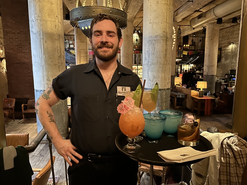 A server holding a tray of colourful cocktails inside Hotel Emma’s historic interior with exposed columns.