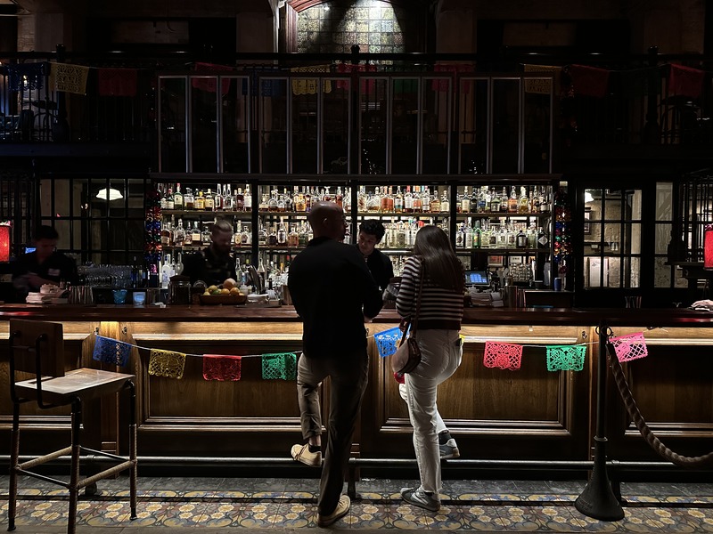 Guests standing at a historic bar inside Hotel Emma, with a backlit display of bottles and warm ambient lighting.