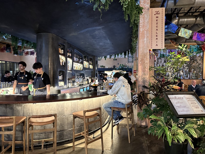 Interior of a mezcaleria at Pullman Market with bartenders preparing drinks and guests seated along a curved bar.