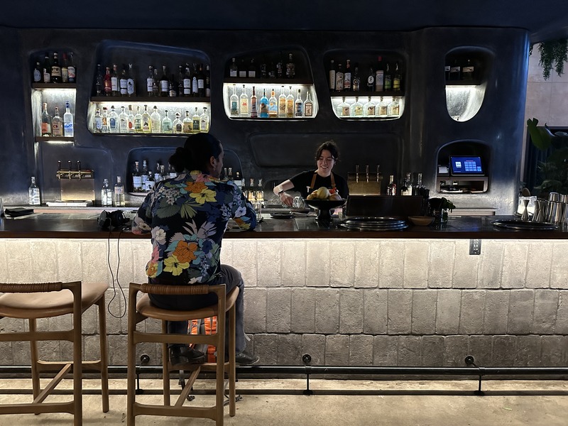 A guest seated at a softly lit mezcal bar with shelves of agave spirits displayed behind the bartender.