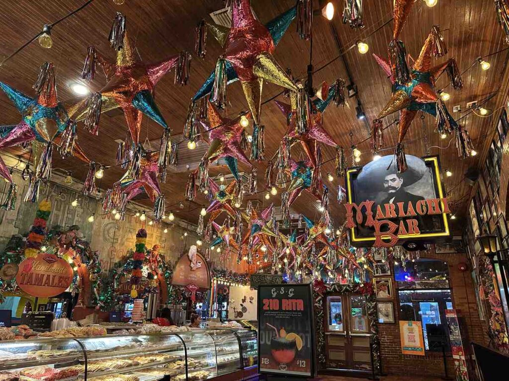 Colourful interior of Mi Tierra Café featuring hanging piñatas, string lights, a pastry counter filled with pan dulce, and a sign for the Mariachi Bar
