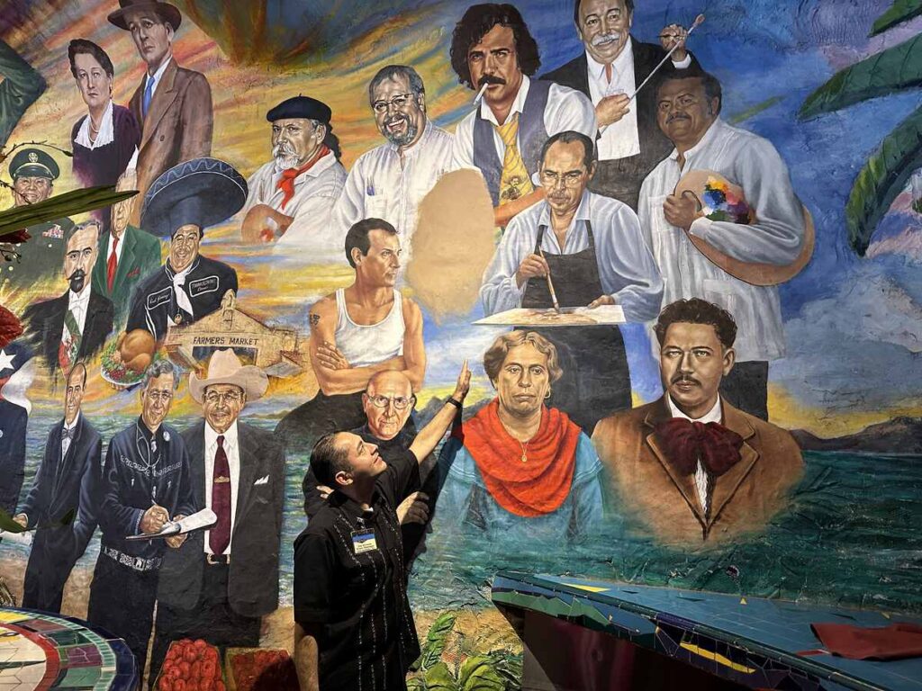 Floor manager pointing at a section of a large mural featuring painted portraits of cultural figures and artists
