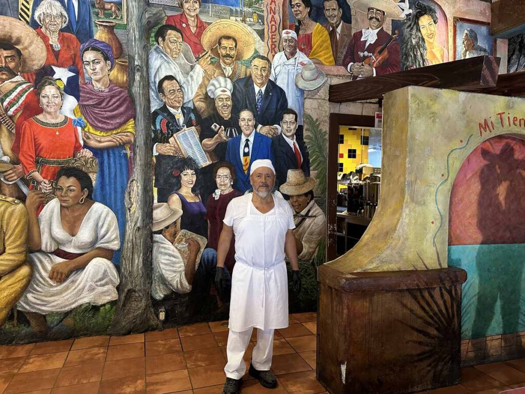 Male baker in white uniform standing proudly in front of a colourful mural inside Mi Tierra Café