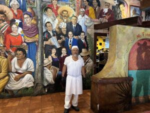 Male baker in white uniform standing proudly in front of a colourful mural inside Mi Tierra Café