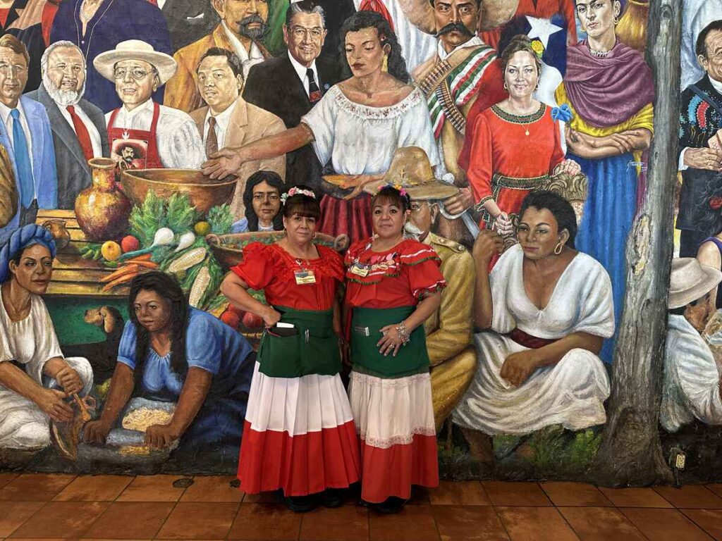 Two female staff members in traditional dress standing in front of a vibrant mural depicting Mexican-American culture and community figures