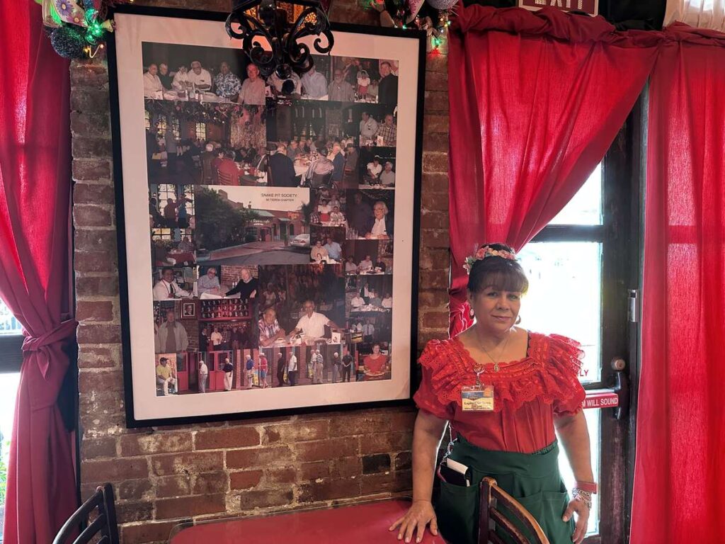Female server in red uniform standing beside a framed collage of photos showing groups of men gathered over decade